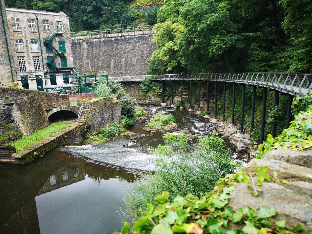 Picture shows a curved bridge over rushing water leading to a building.